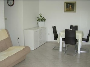 A living room with a sofa, a dining table with chairs, a white cabinet, a potted plant, a clock, and a framed picture on the wall. - Apartments Vesna (Slivno)