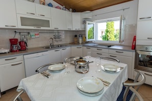 A kitchen with white cabinets, a Bosch coffee maker, a window with a view of greenery, and a dining table set with plates and wine glasses. - Apartments Vesna (Komarna)