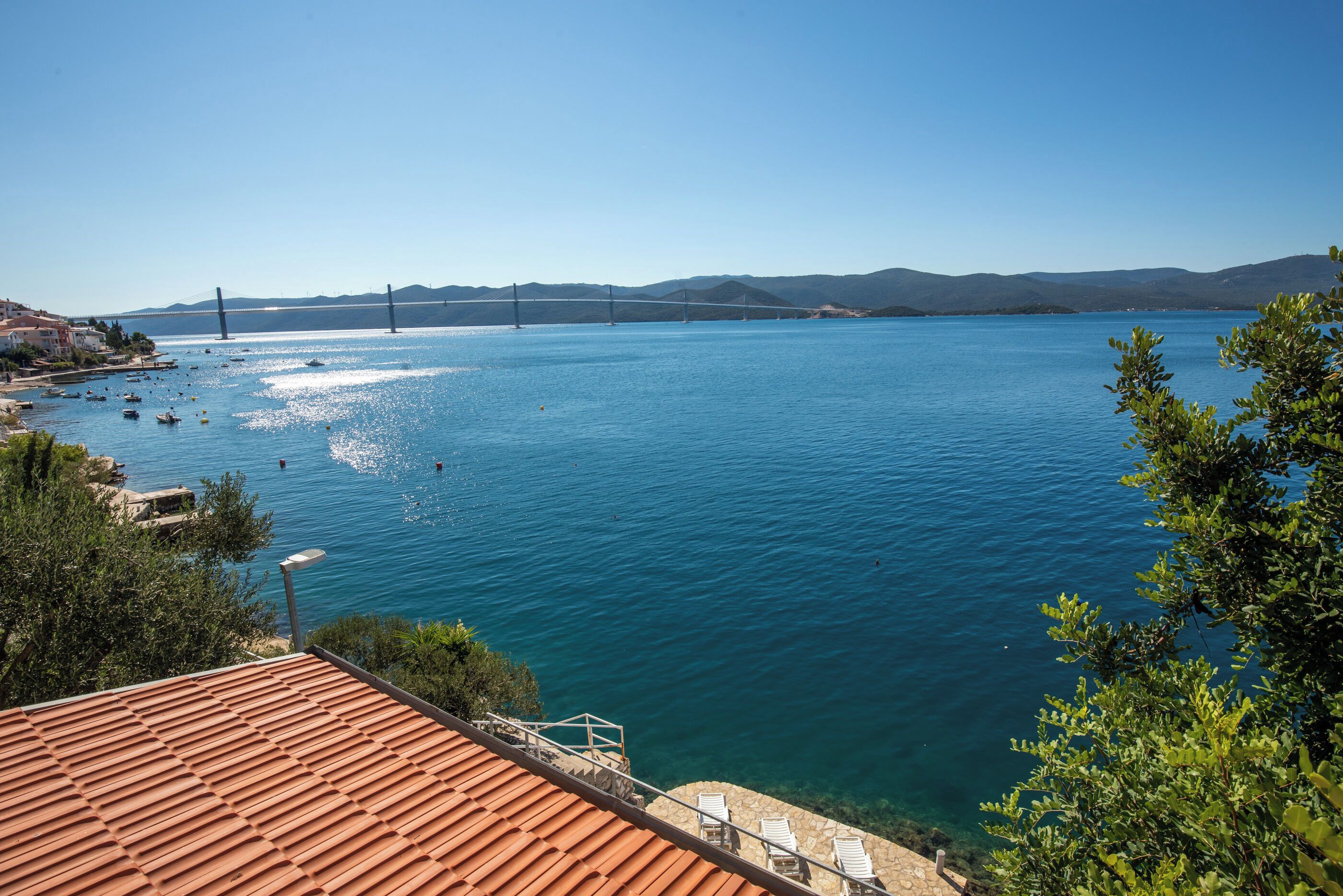 A coastal view with a clear blue sea, a bridge in the distance, and a building with a tiled roof.