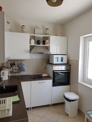 A compact kitchen with white cabinetry, a microwave, an oven, and a trash bin. - Apartments Vesna (Slivno)