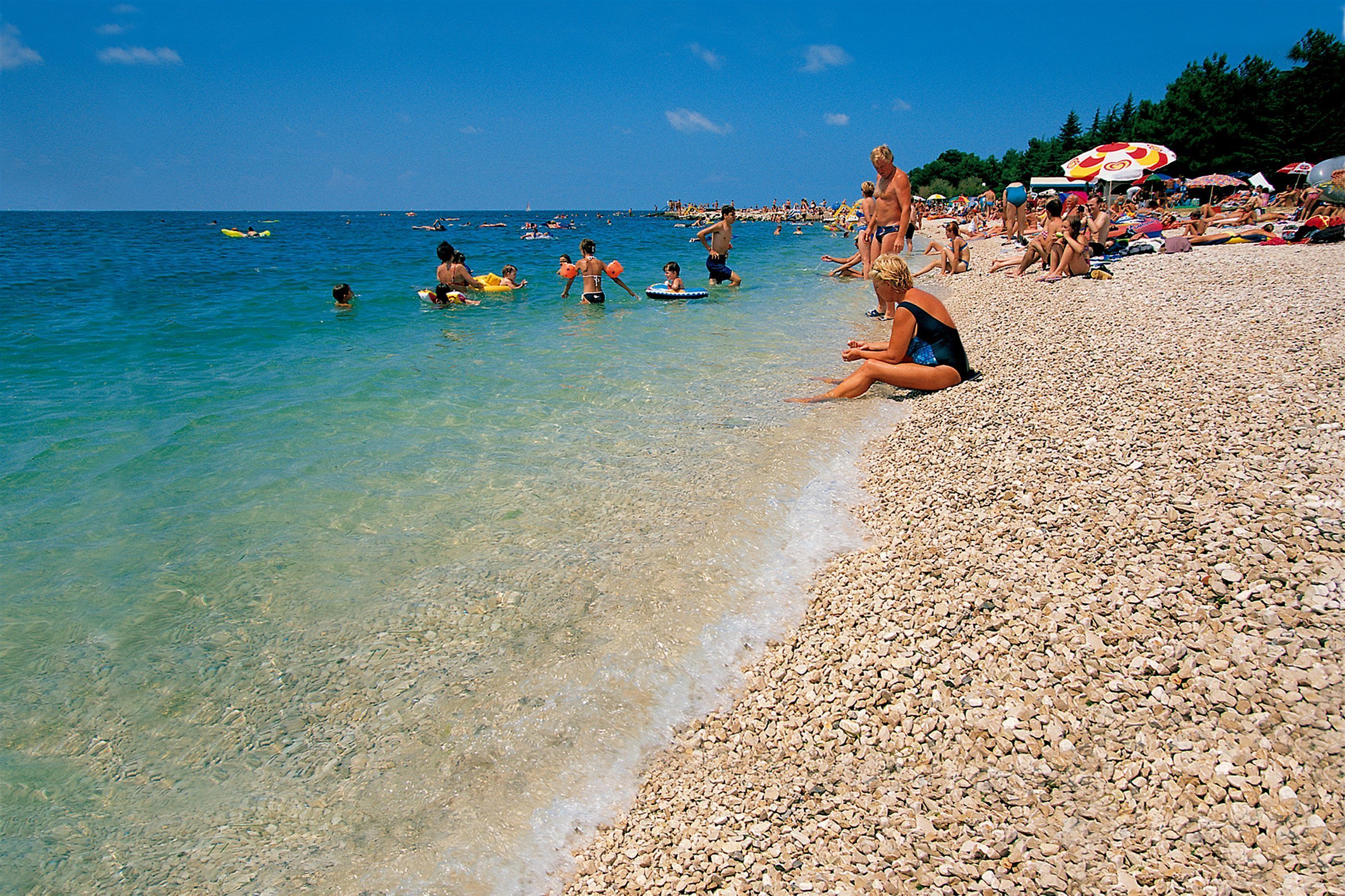 Una playa de guijarros con agua cristalina y gente nadando y tomando el sol.