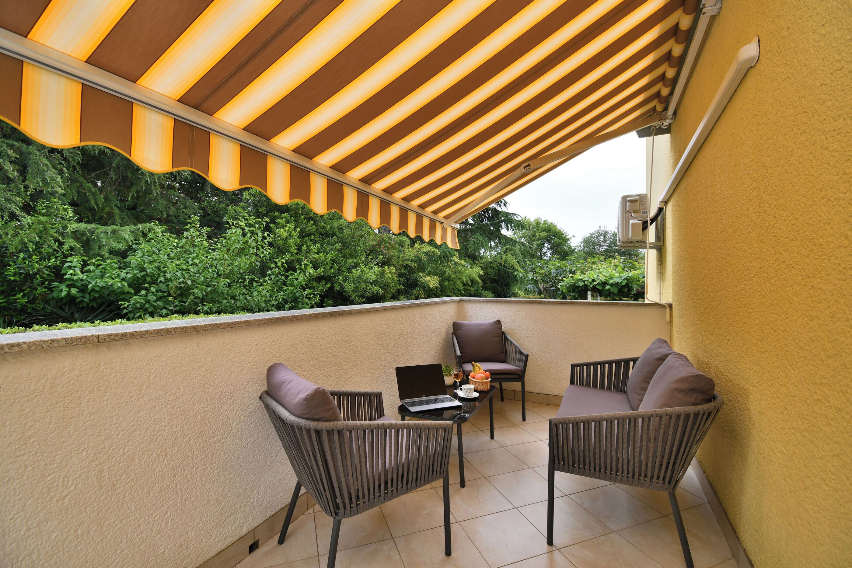 Un balcon agrémenté d’un auvent à rayures, deux fauteuils en osier, une petite table avec un ordinateur portable et un panier de fruits, et une vue sur la verdure.