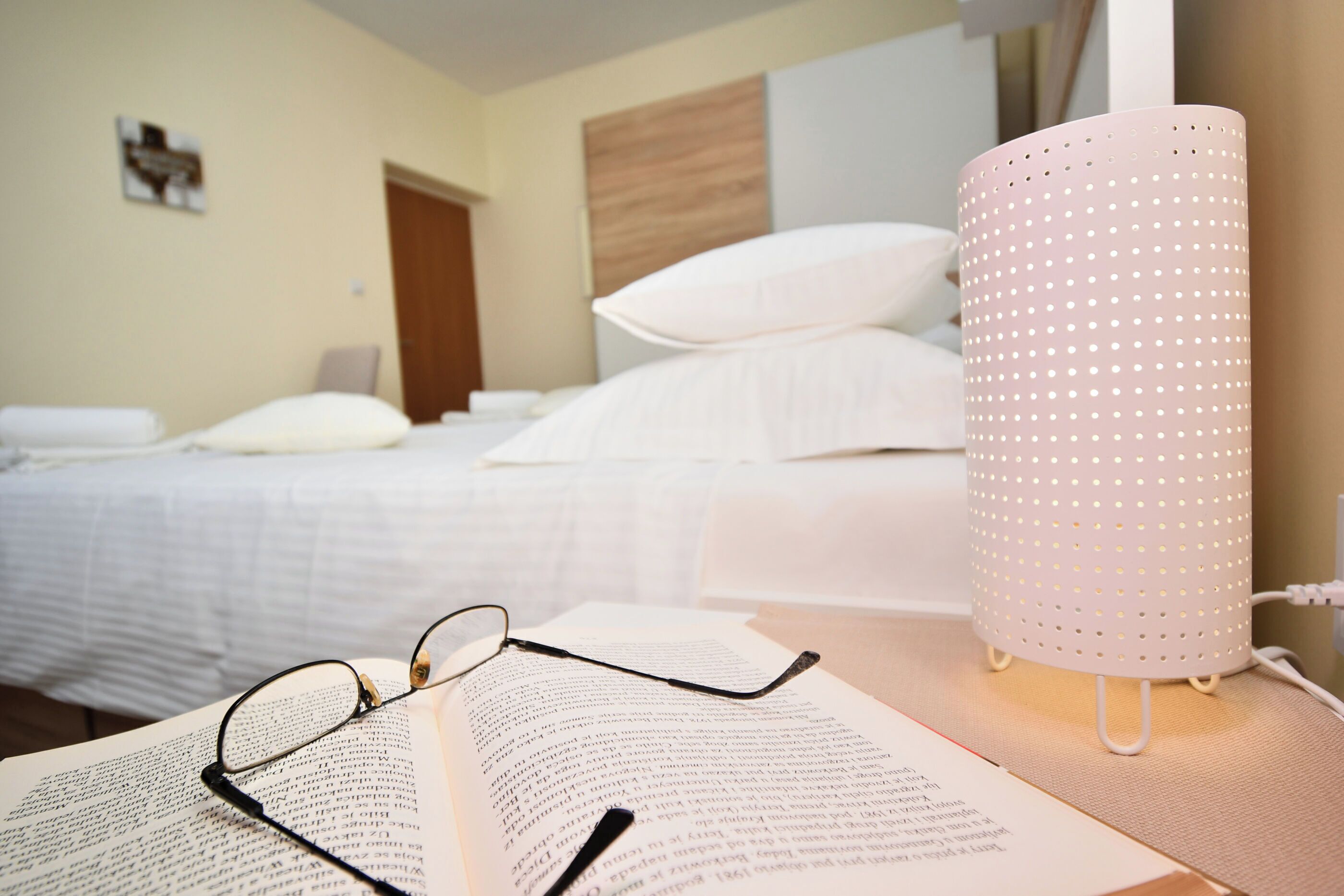 A hotel room with a bed, a bedside table with a lamp, and an open book with glasses on it.