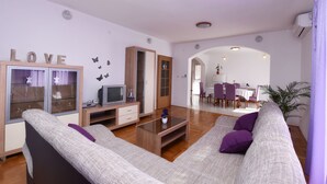 Una sala de estar moderna con un sofá, televisor y un mueble. Se observa un comedor al fondo.
