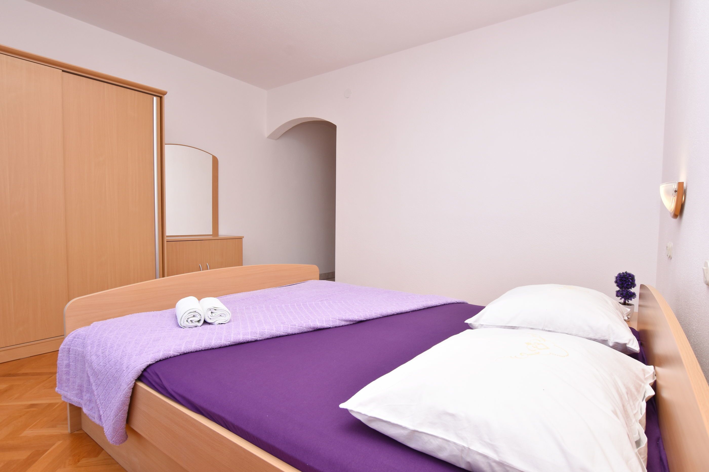 A neatly made bed with a purple bedspread and white pillows, a wooden wardrobe, and a small potted plant on the nightstand.