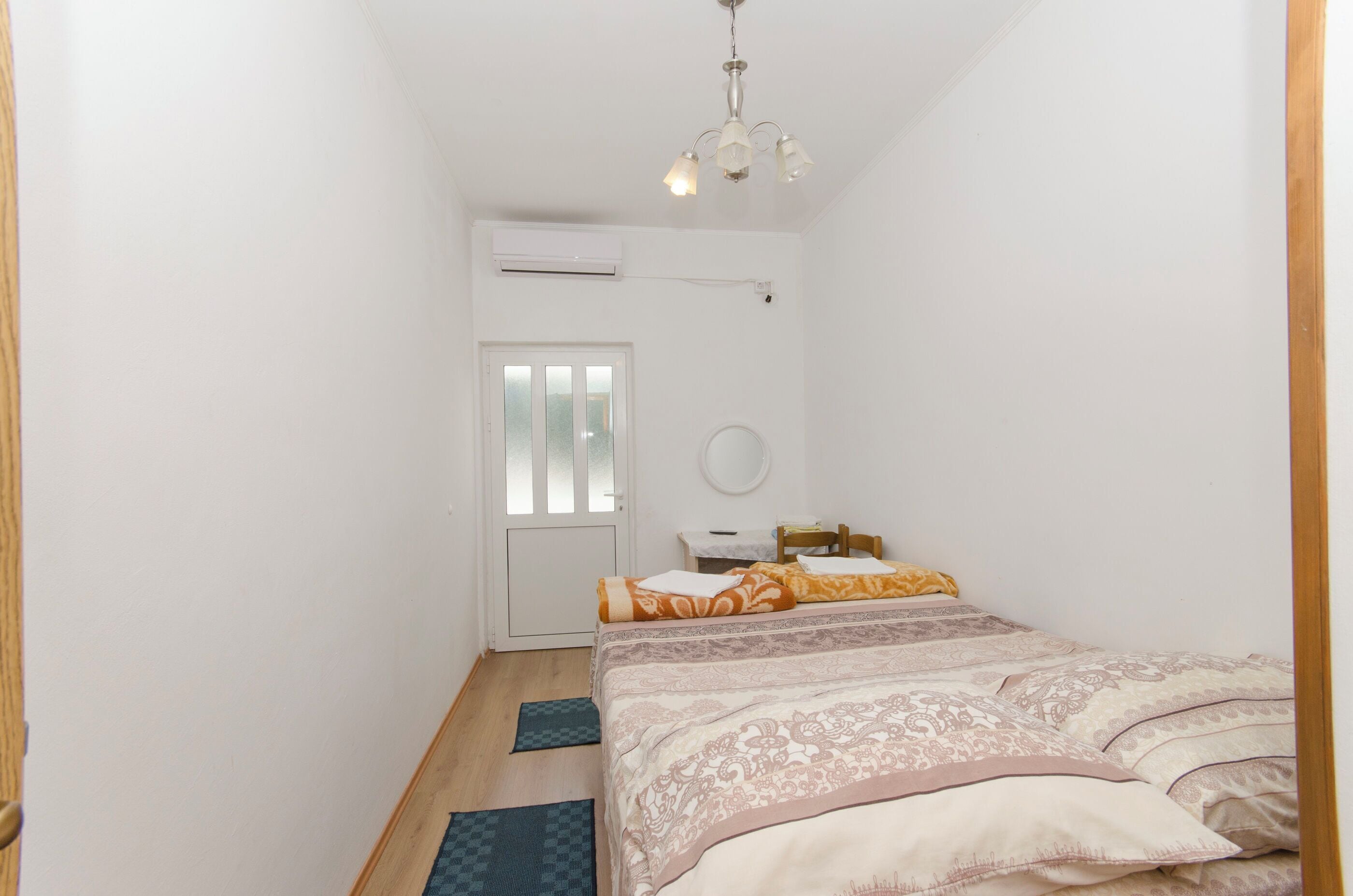 A bedroom with a bed, a desk, and a chair. There is a door and an air conditioning unit on the wall.