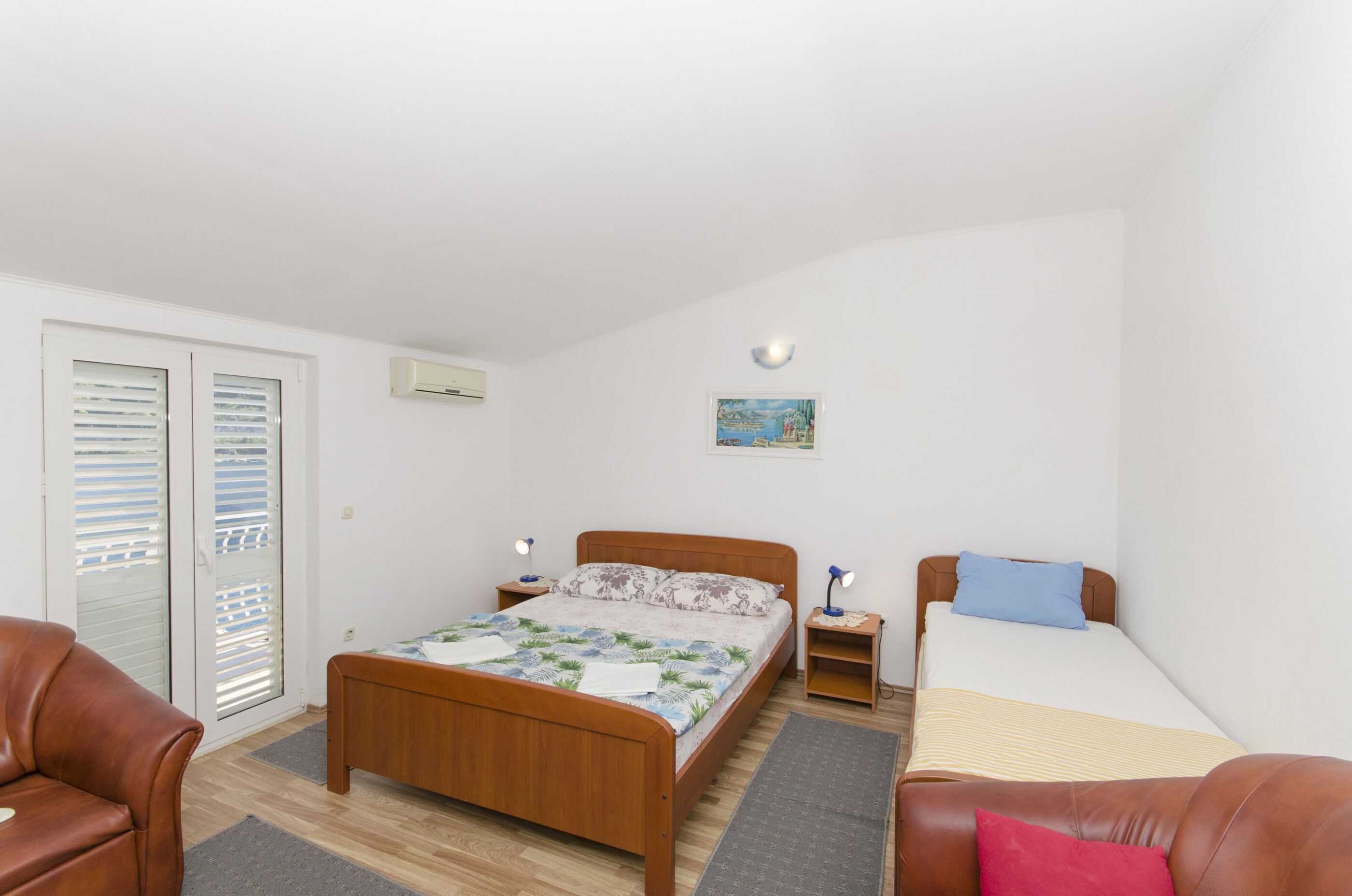Un dormitorio con cama de madera, un sofá, una mesita pequeña y una mesita de noche. Hay una ventana con persianas, un aire acondicionado instalado en la pared y un cuadro en la pared.