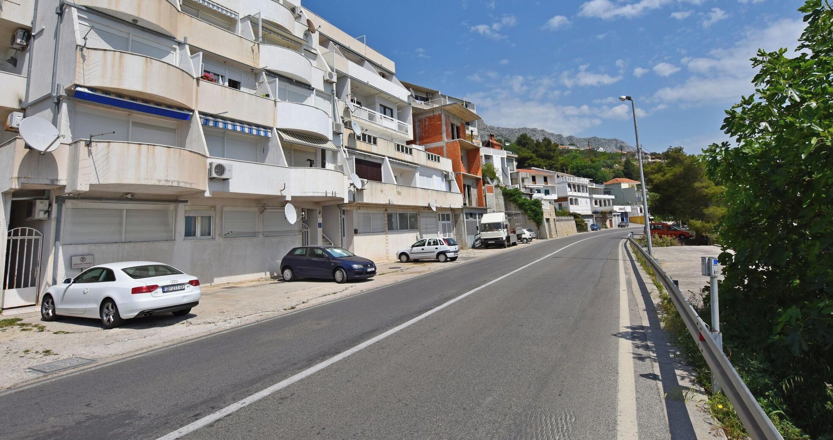A street lined with multi-story residential buildings, parked cars, and a clear sky.