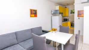 Un salon moderne avec un canapé gris, une table blanche et des chaises assorties. Une petite table avec un vase de fleurs est placée au centre. La cuisine, visible à l’arrière-plan, est dotée de meubles jaunes et d’appareils électroménagers blancs.