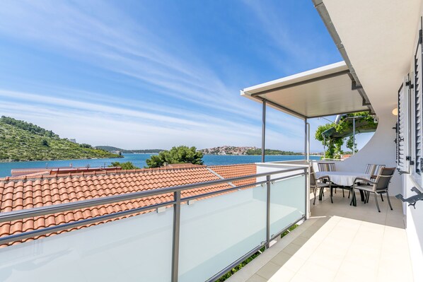 A balcony with a glass railing overlooking a coastal area with terracotta tiles and a table with chairs. - Apartments Vincic (Rogoznica)