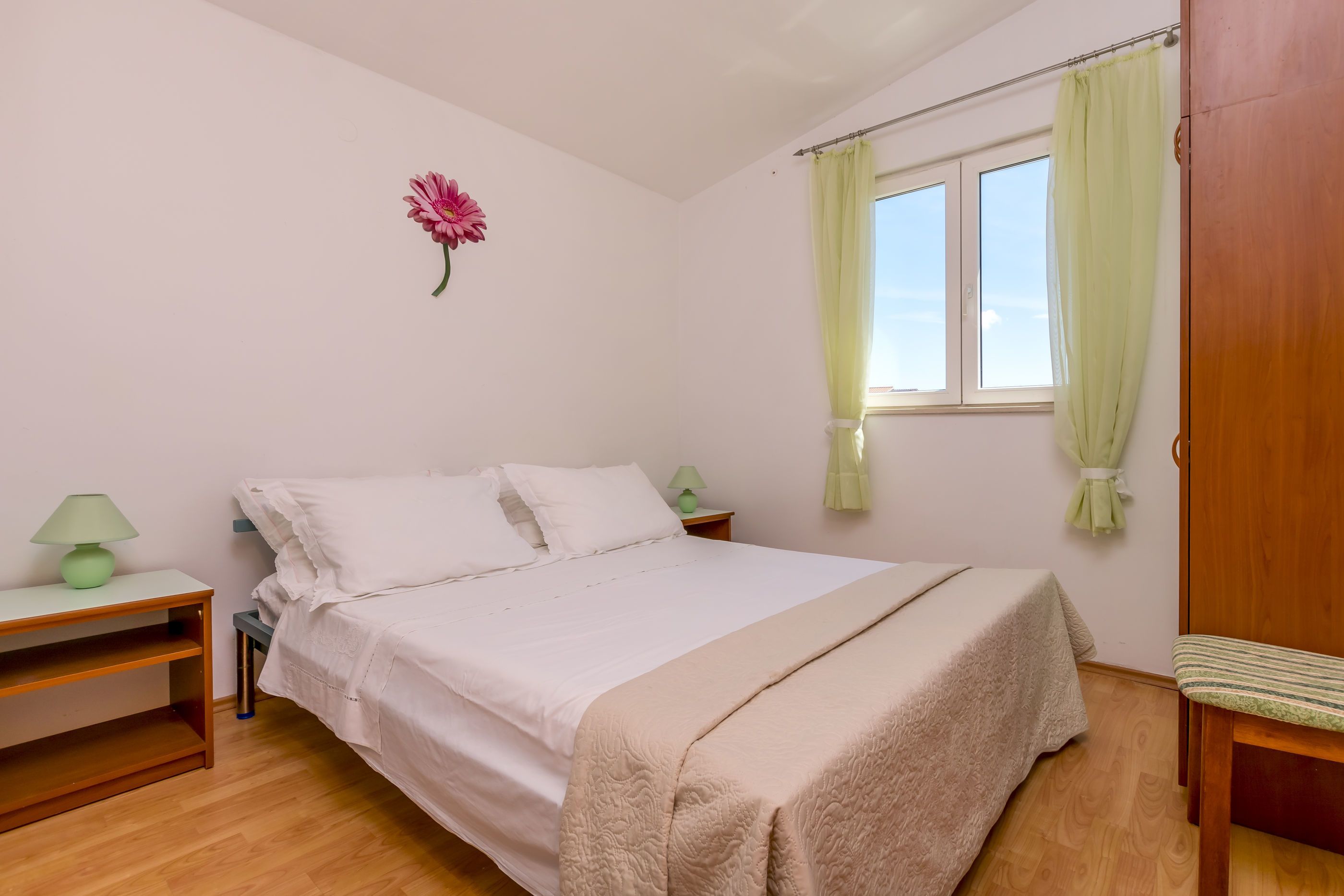 Un dormitorio con cama, mesita de noche, lámpara, ventana con cortinas y una silla de madera.