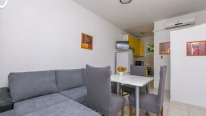 Un salon moderne avec un canapé gris, une table blanche et deux fauteuils gris. On aperçoit à l’arrière-plan un coin cuisine avec des placards jaunes et un réfrigérateur blanc.