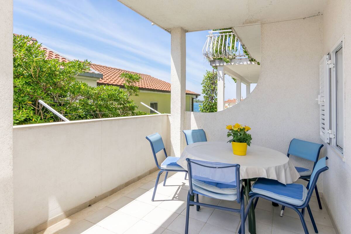 A balcony with a round table and blue chairs, a potted plant, and a view of buildings and trees.