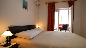 A hotel room with a bed, a nightstand with a lamp, a window with red curtains, and a view of an outdoor area with chairs. - Apartments and Room Janja (Primosten)