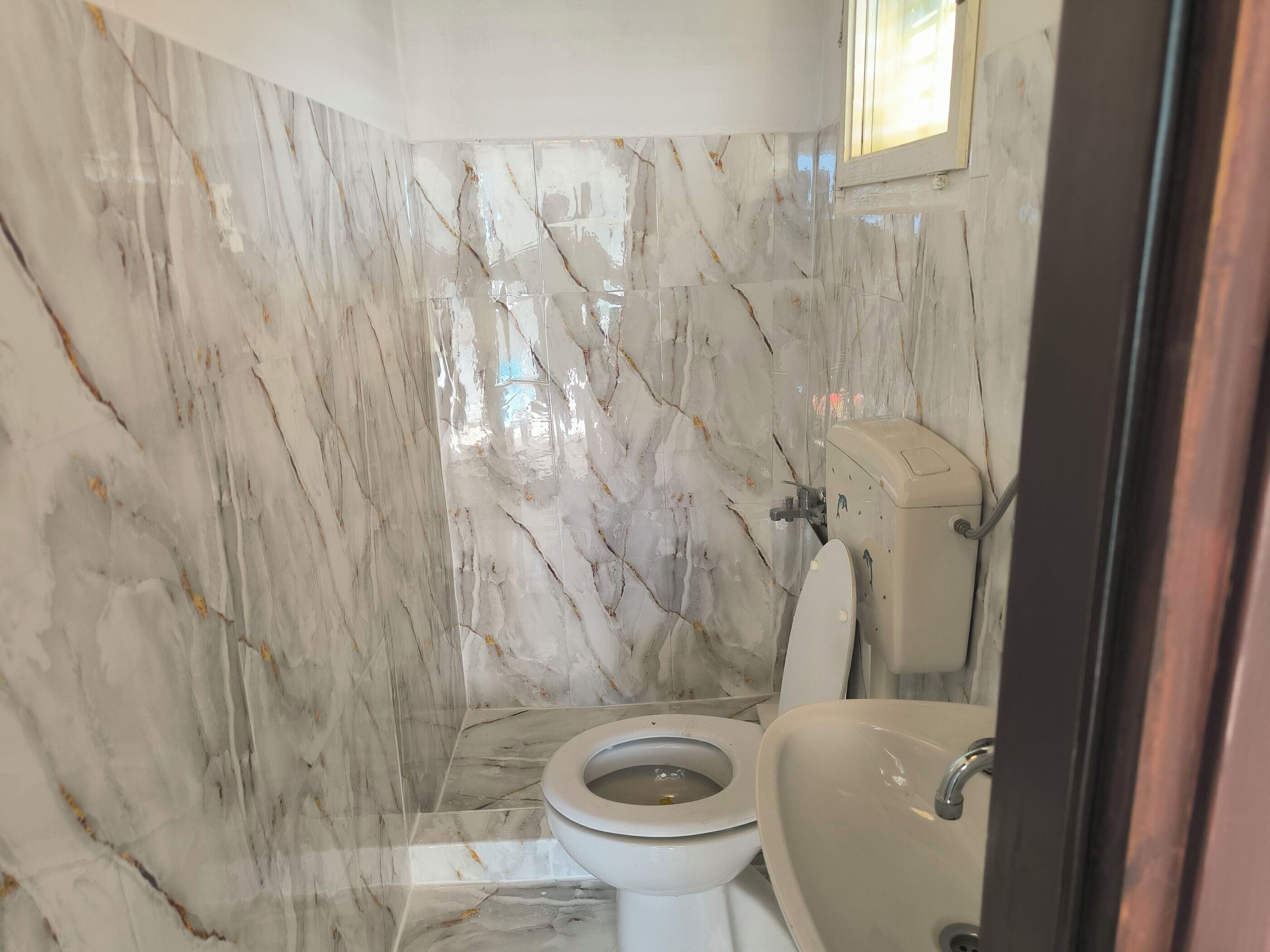 A bathroom with a toilet, sink, and shower area, featuring marble walls and a window.
