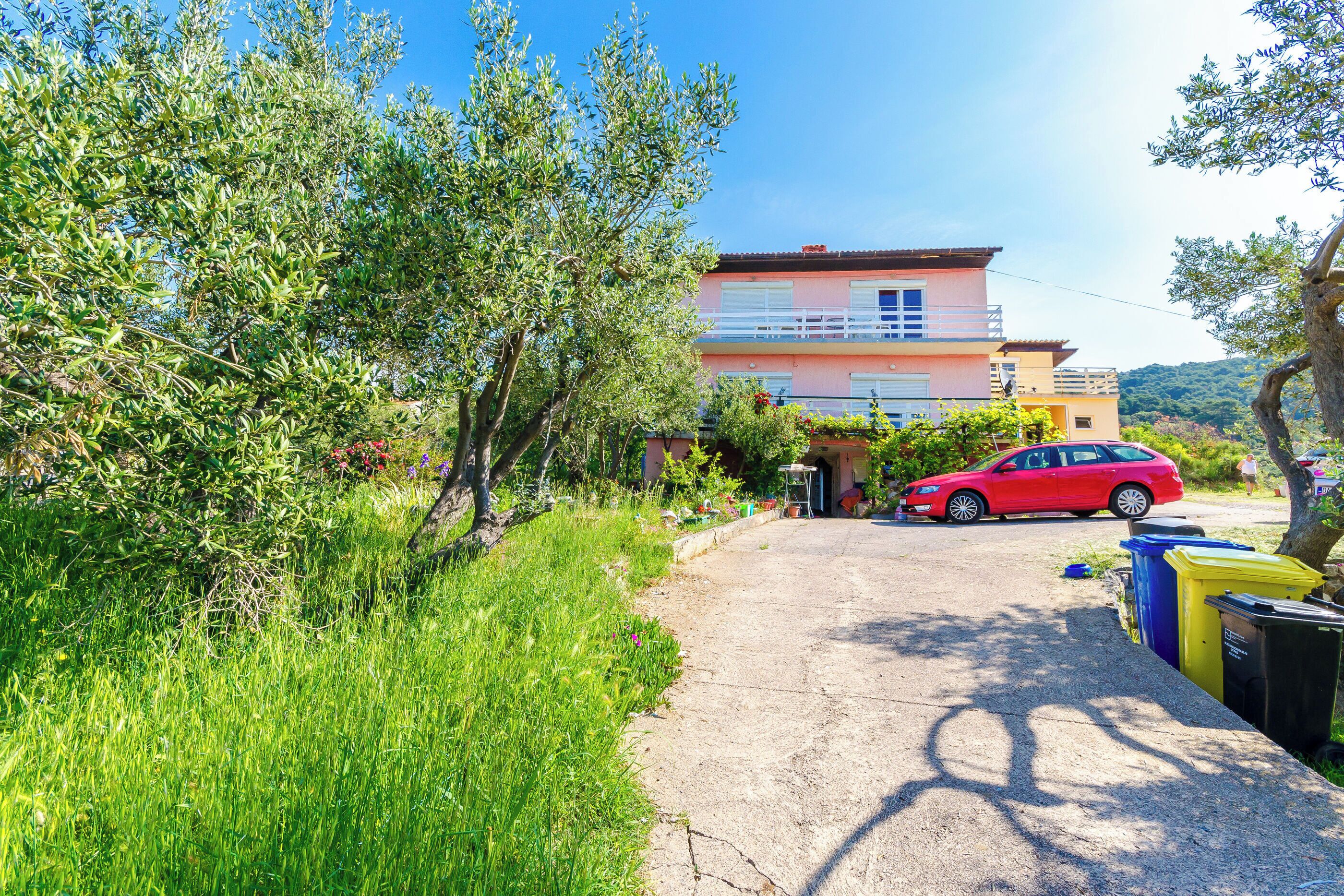A residential area with a red car parked in front of a house with a balcony. There are trash bins along the side of the road.