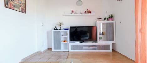 A modern living room with a flat-screen TV, a white entertainment unit, and a dining table with a vase of flowers.