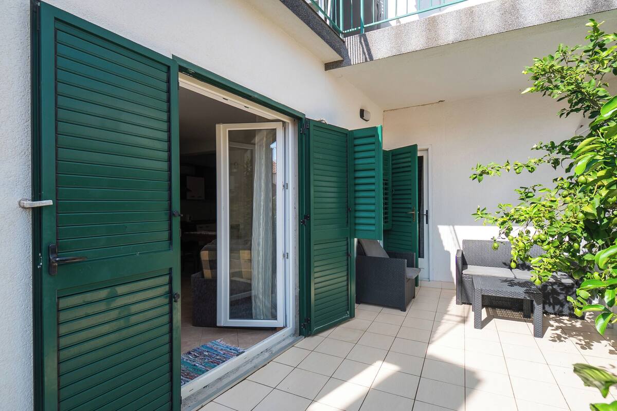 A residential balcony with green shutters, a sliding glass door, outdoor furniture, and a potted plant.