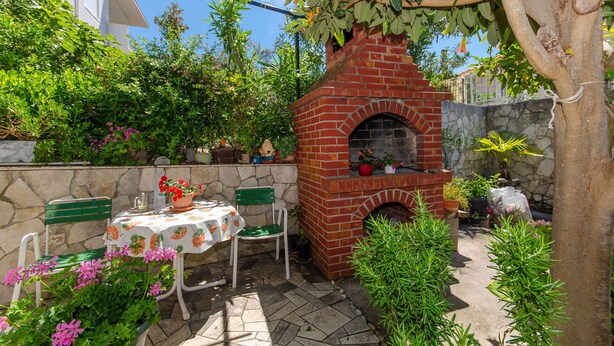 Ein gemauerter Außenofen, ein Tisch mit blumigem Tischläufer und Stühle in einem Garten.