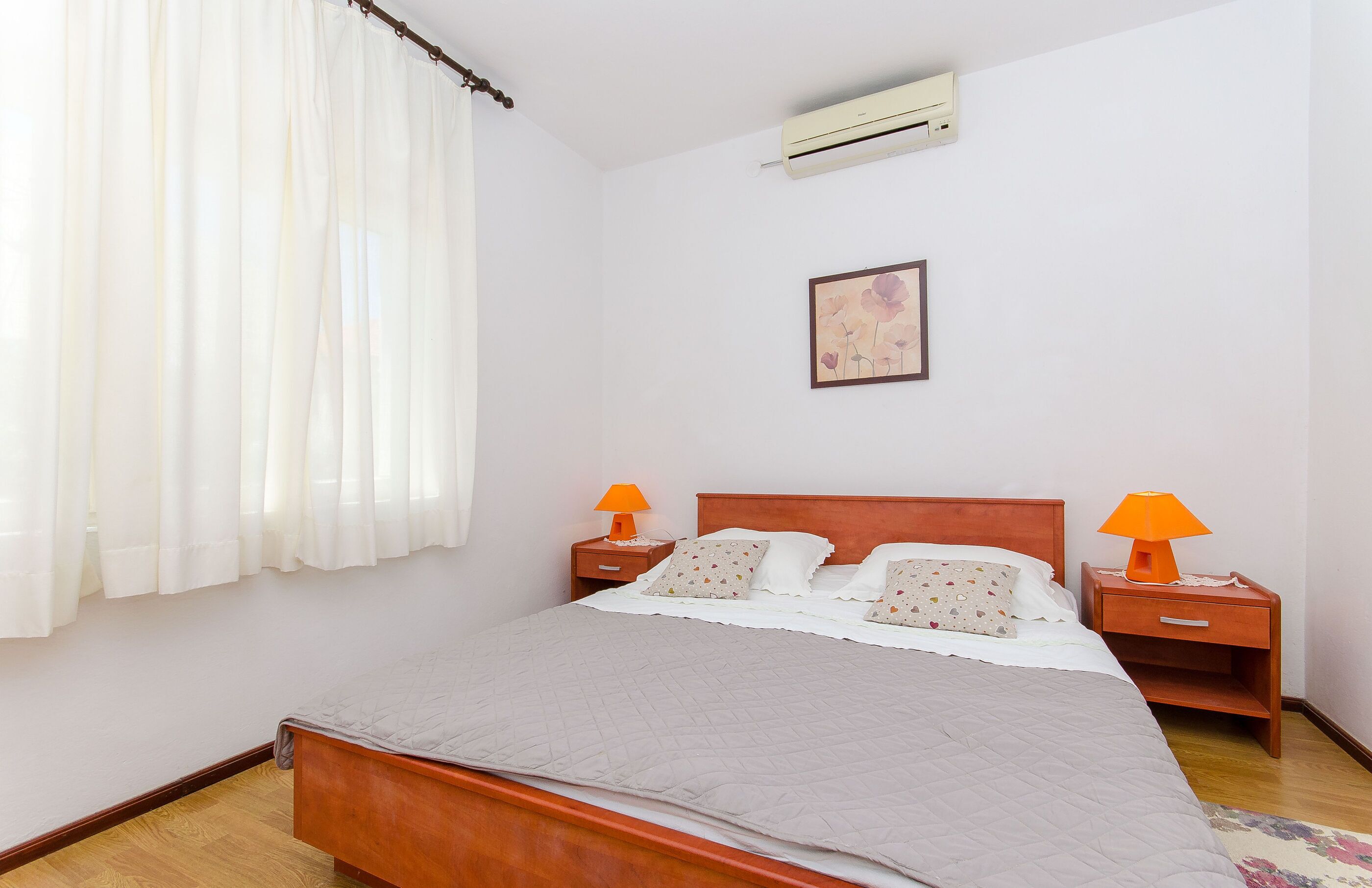 A neatly made bed with a wooden headboard, two bedside tables with lamps, a wall-mounted air conditioner, and a framed picture on the wall.