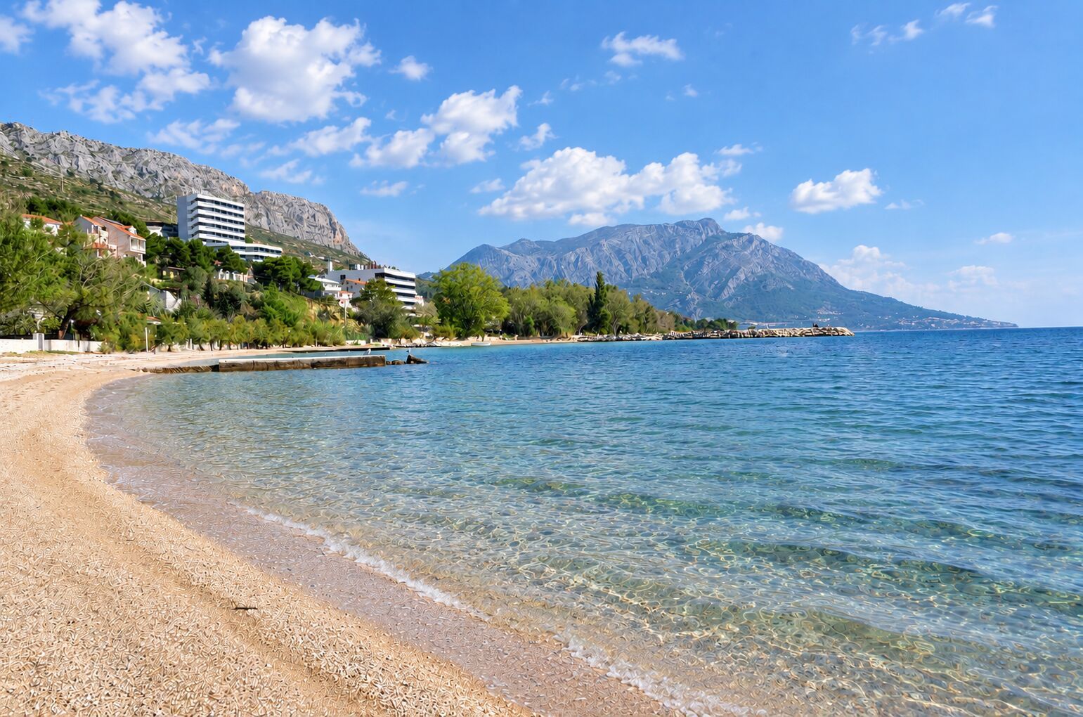 A beach with clear water, a rocky shoreline, and buildings in the background.