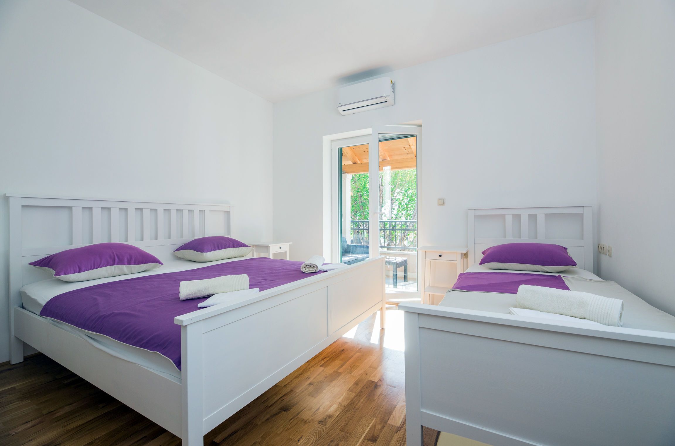 A bedroom with two beds, each with a white headboard and purple bedding. There is a window with a view of greenery outside.