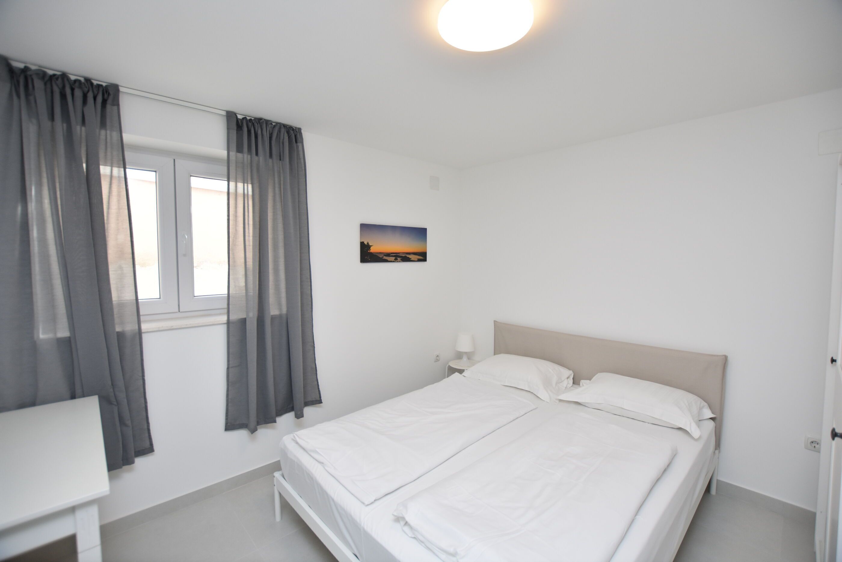 A bedroom with a bed, a bedside table, a nightstand, a window with curtains, and a wall-mounted picture.