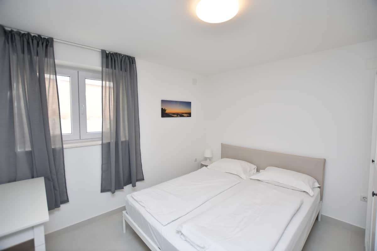 A bedroom with a bed, a bedside table, a nightstand, a window with curtains, and a wall-mounted picture.