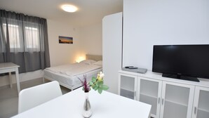 Ett hotellrum med en säng, en tv, ett skrivbord, en stol, ett vitt skåp och en vas med blommor.