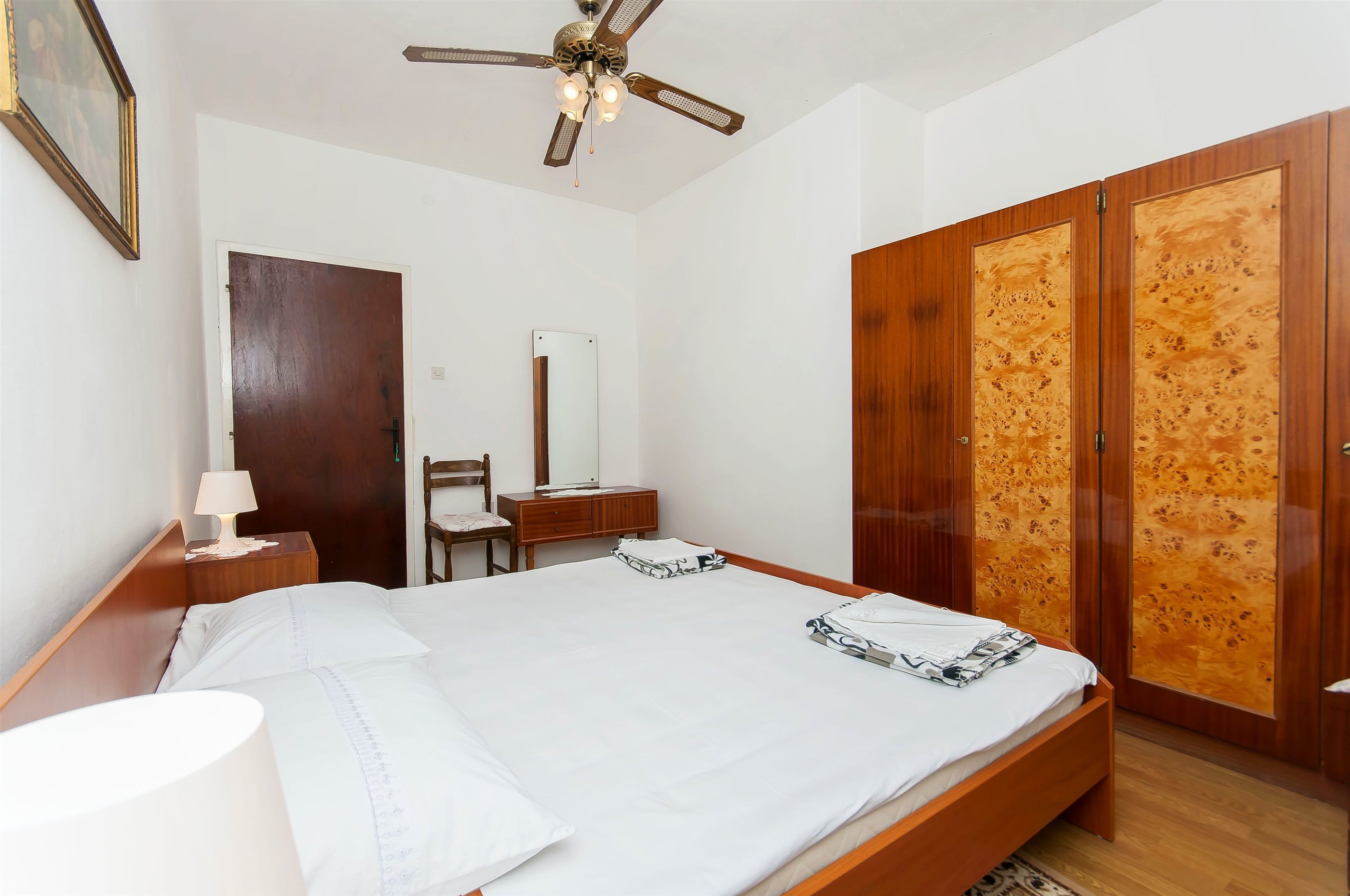 A neatly arranged bedroom with a bed, bedside table, lamp, wooden wardrobe, and a ceiling fan.
