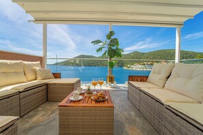 Un balcón con mobiliario de mimbre, una mesa con alimentos para el desayuno y vistas al mar y las colinas.