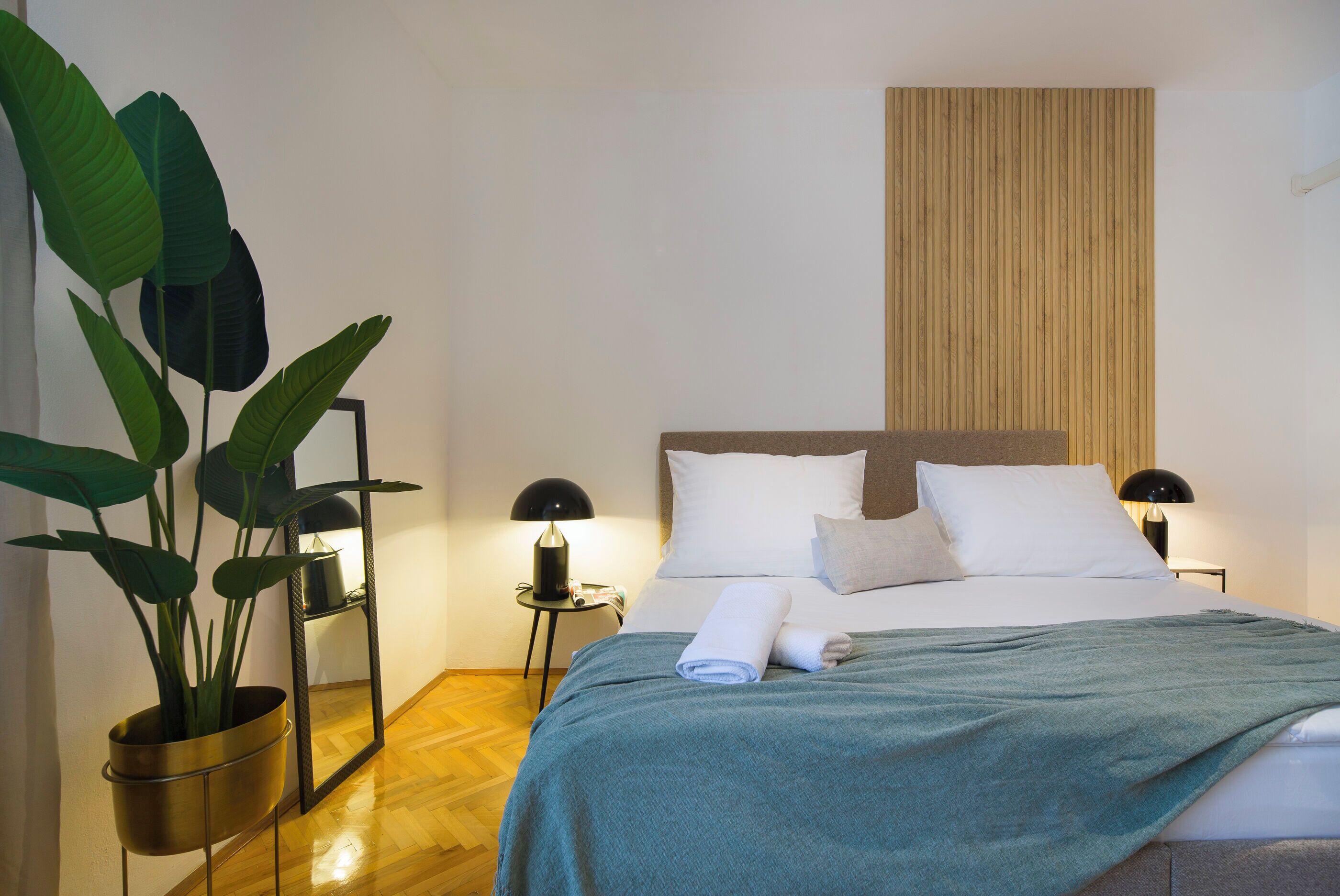 Un lit bien fait, avec du linge de lit blanc et une couverture turquoise, une plante en pot posée sur un support et un miroir sur une table d’appoint.