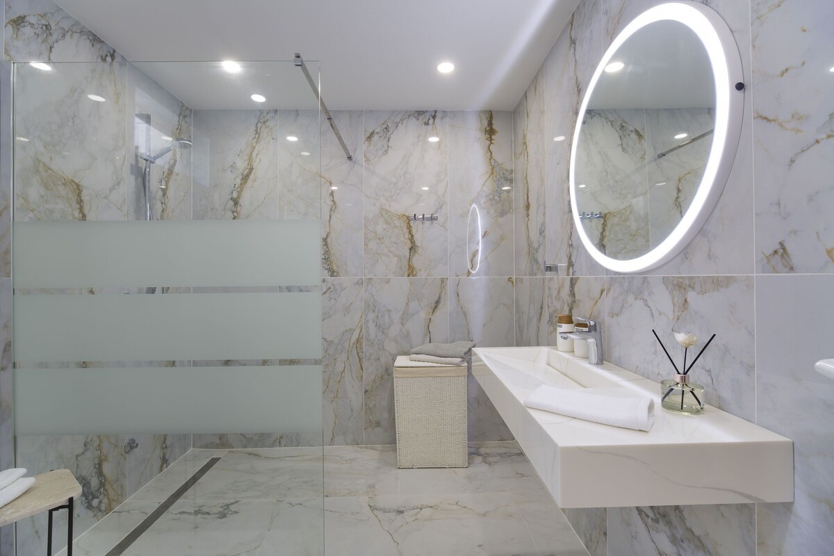 A modern bathroom with a glass-enclosed shower, a white vanity with a mirror, and marble walls.