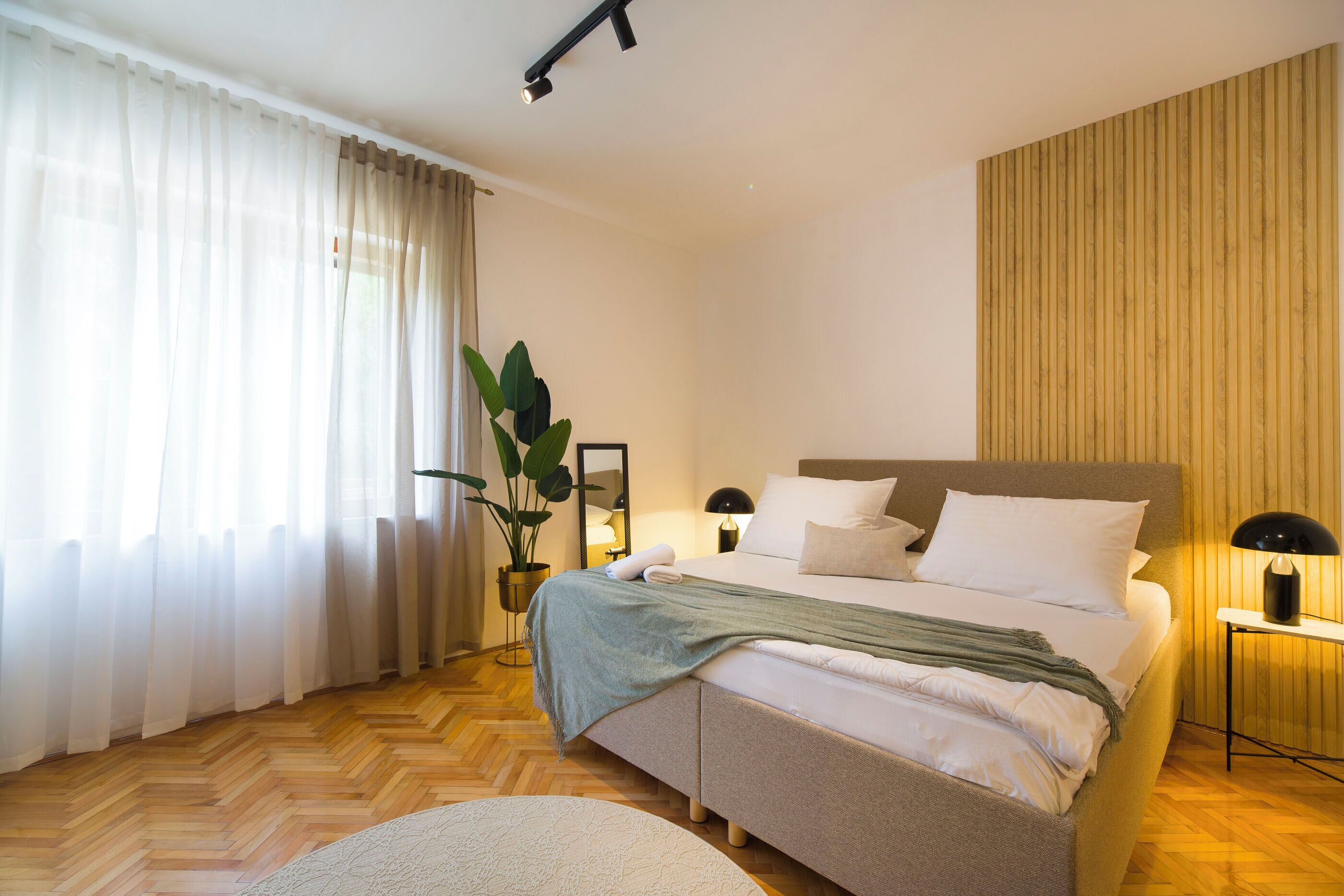 A modern bedroom with a large bed, a bedside table with a lamp, a potted plant, and a wooden panel wall.