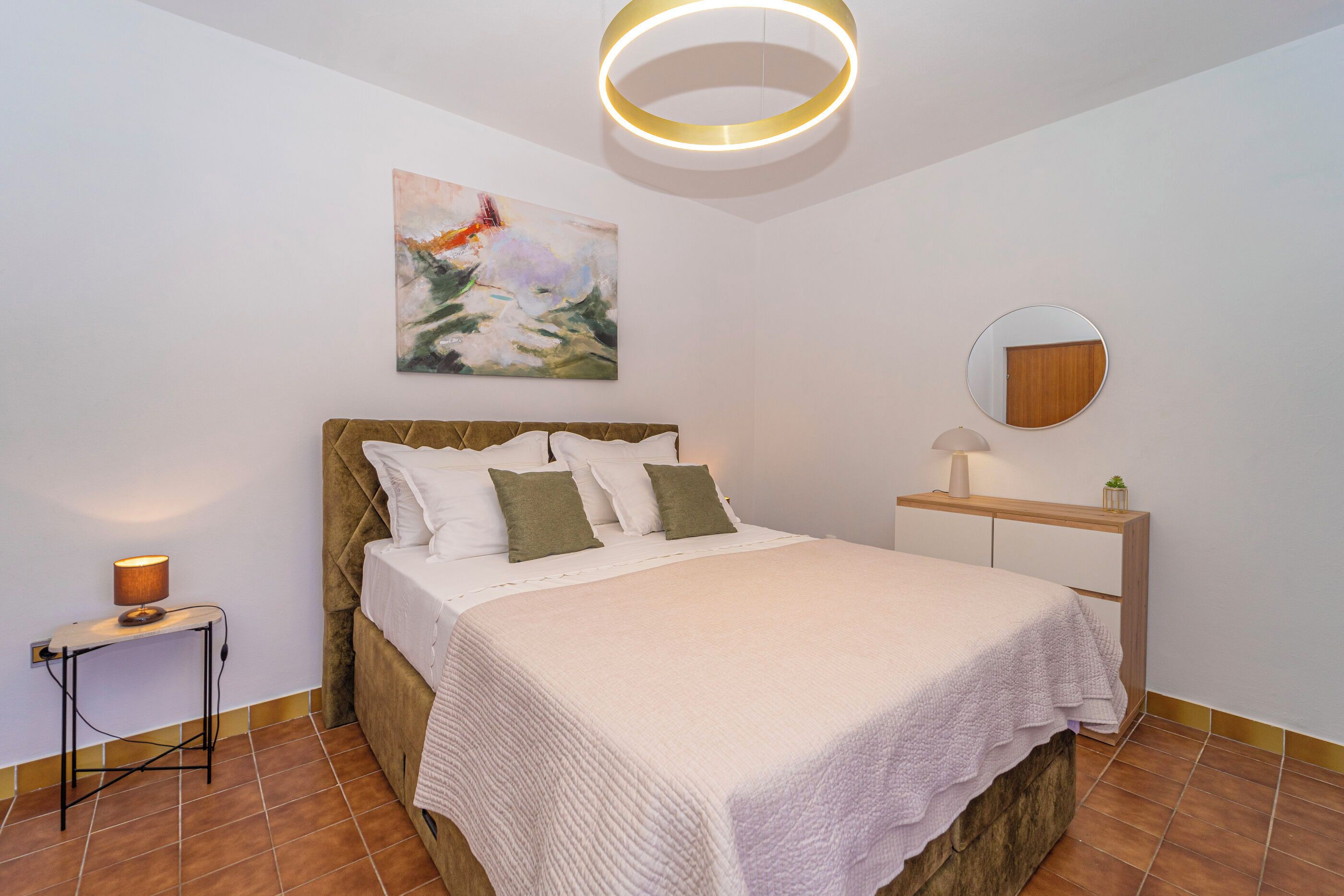 A bedroom with a bed, a bedside table, a lamp, a wall-mounted mirror, and a painting.