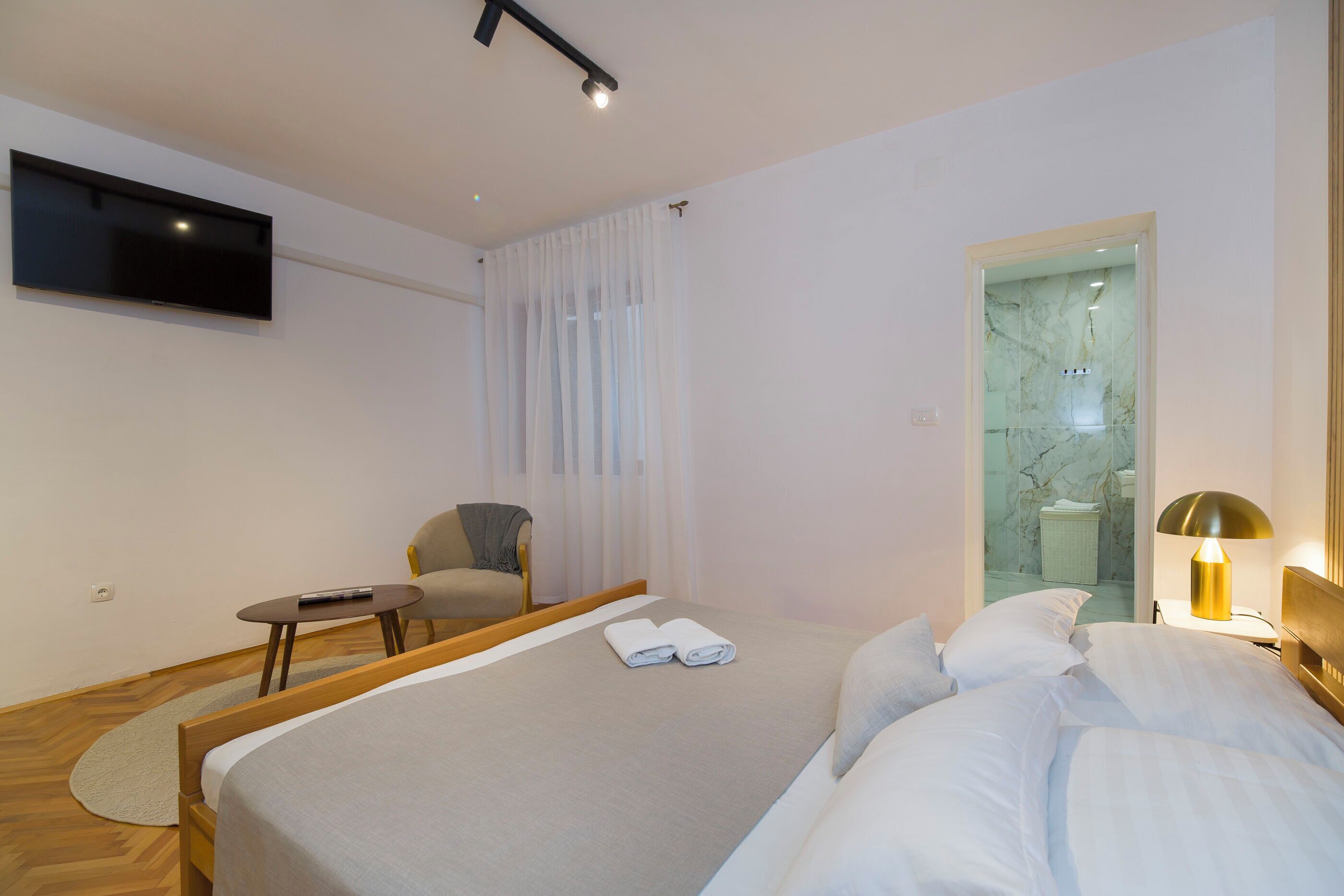 Habitación de hotel moderna con una cama grande, un televisor de pantalla plana en la pared, una mesa redonda pequeña con una silla y un baño visible a través de una puerta de cristal.
