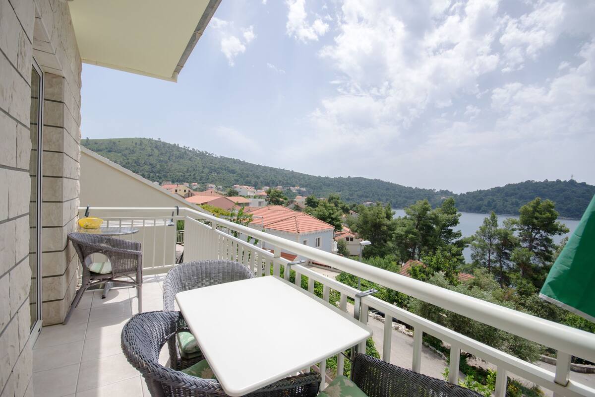 A balcony with wicker furniture, a white table, and a view of a lake and hills.