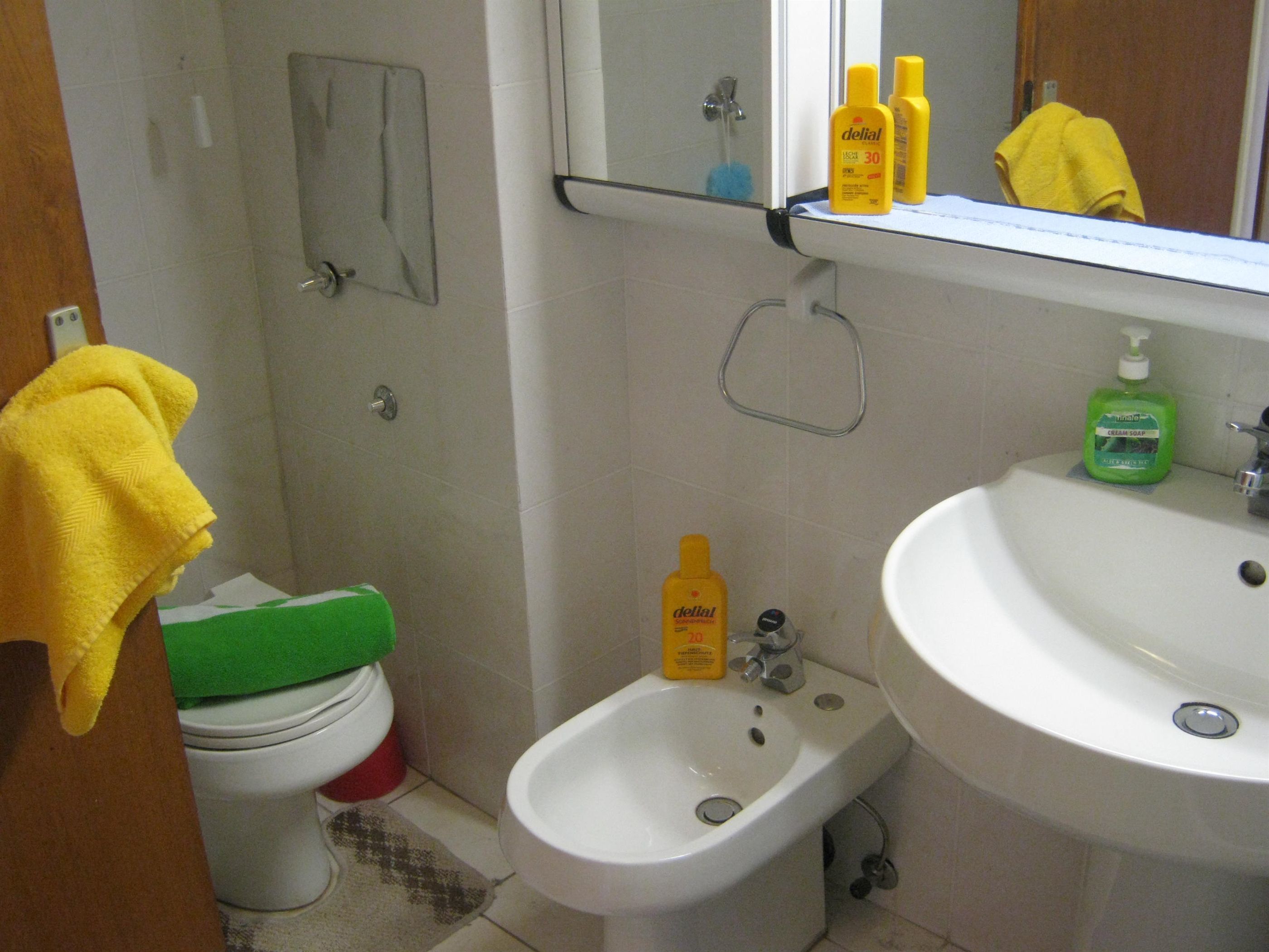 A bathroom with a toilet, bidet, sink, and mirror. There are yellow towels and a green bottle of liquid soap on the sink.