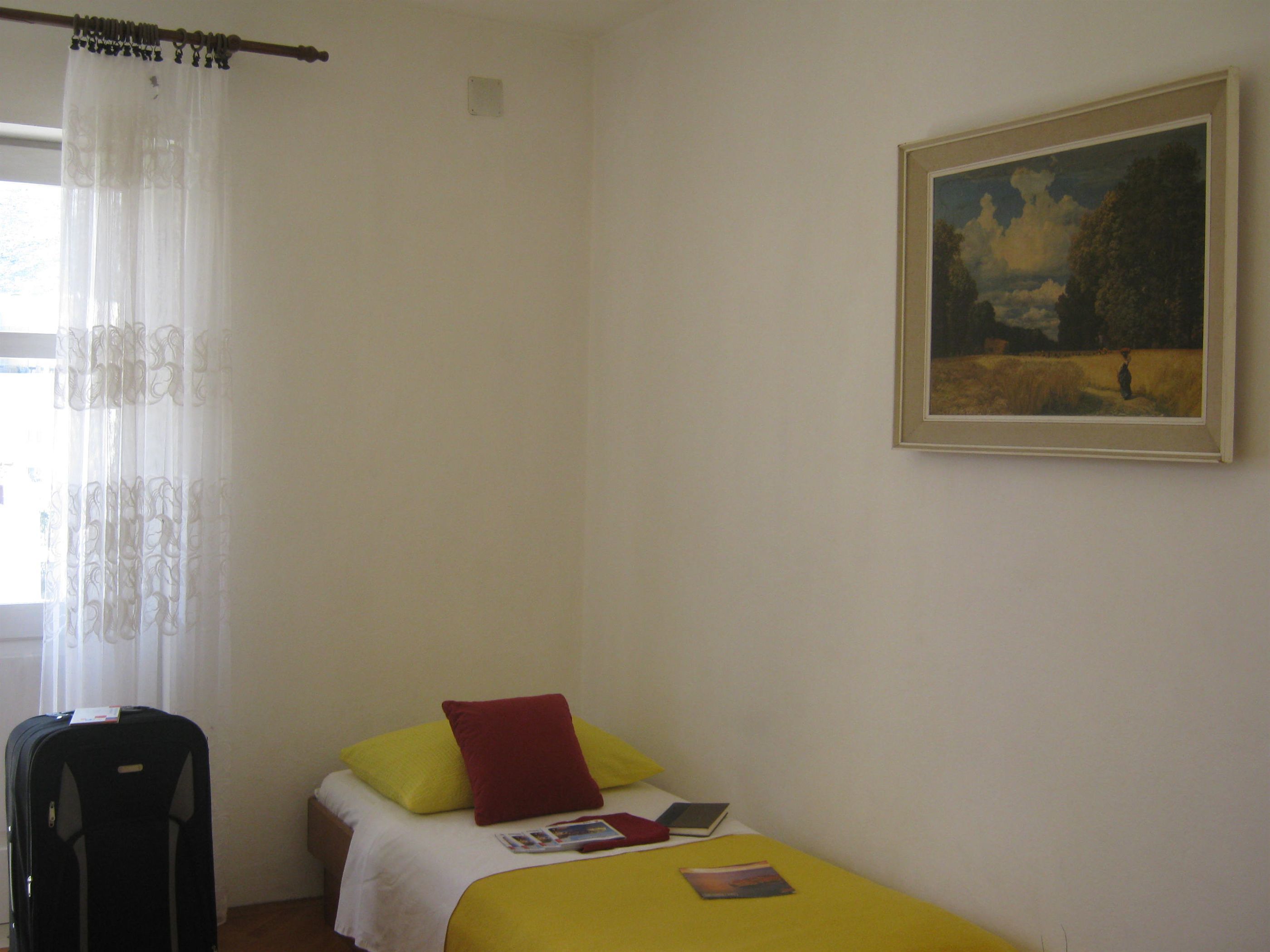A small, simple room with a bed, a chair, a painting, and a window with curtains.