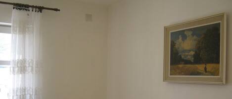 A small, simple room with a bed, a chair, a painting, and a window with curtains.