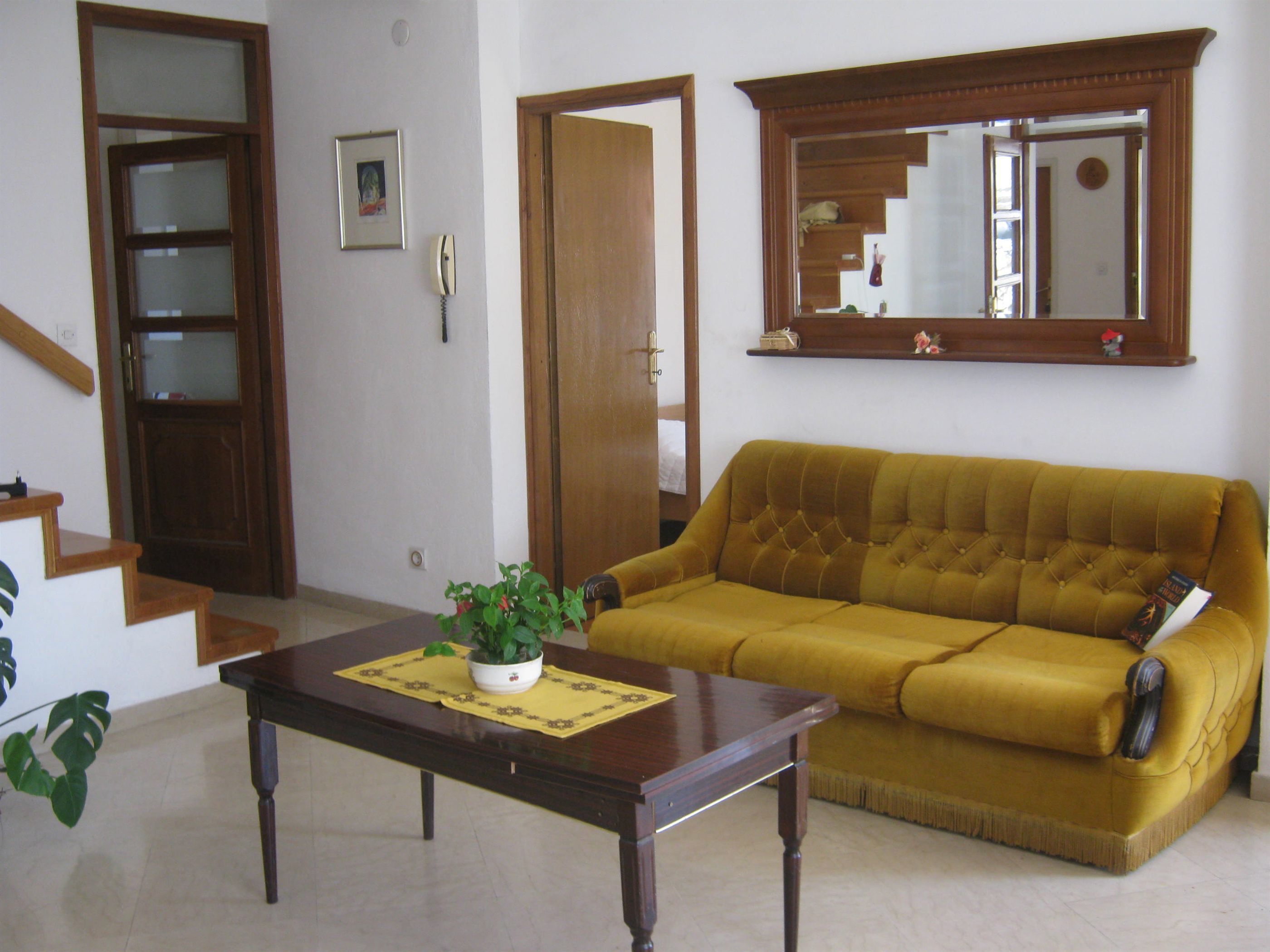 Uma sala de estar com um sofá amarelo, uma mesa de centro de madeira com uma planta, um quadro na parede e uma porta de madeira.