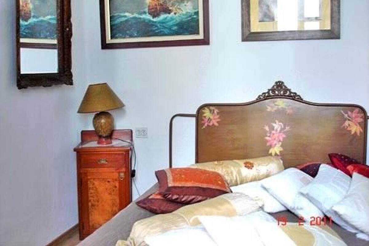 A bedroom with a wooden headboard, a bedside table with a lamp, and a small cabinet. There are three framed pictures on the wall.