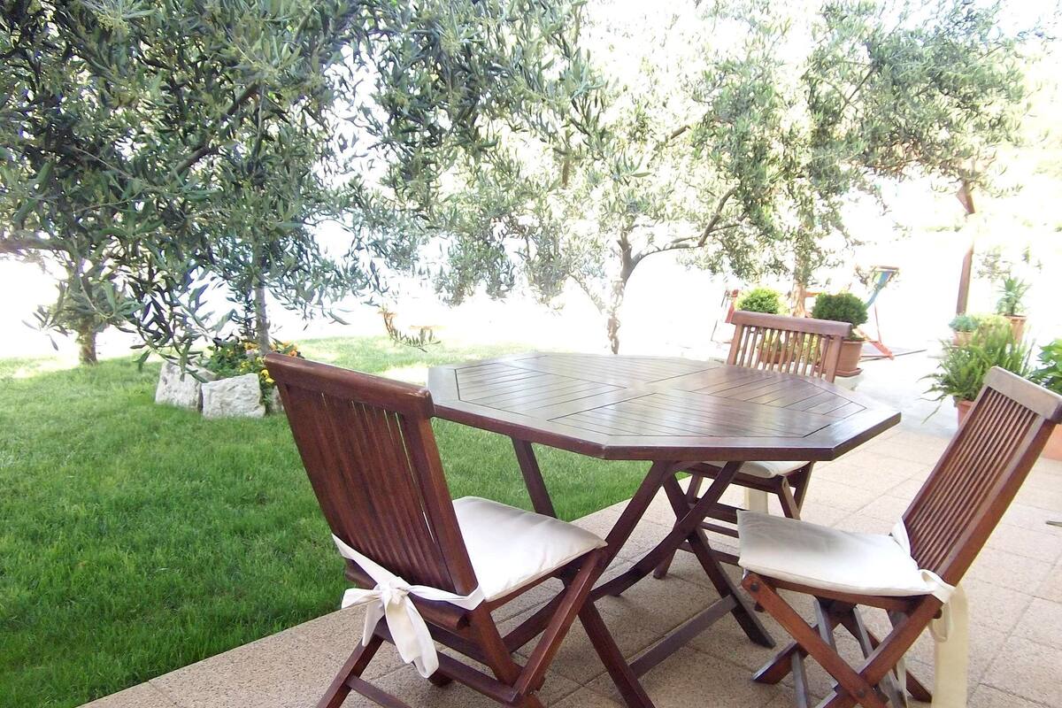 An outdoor patio with a round wooden table and four wooden chairs, surrounded by greenery and potted plants.