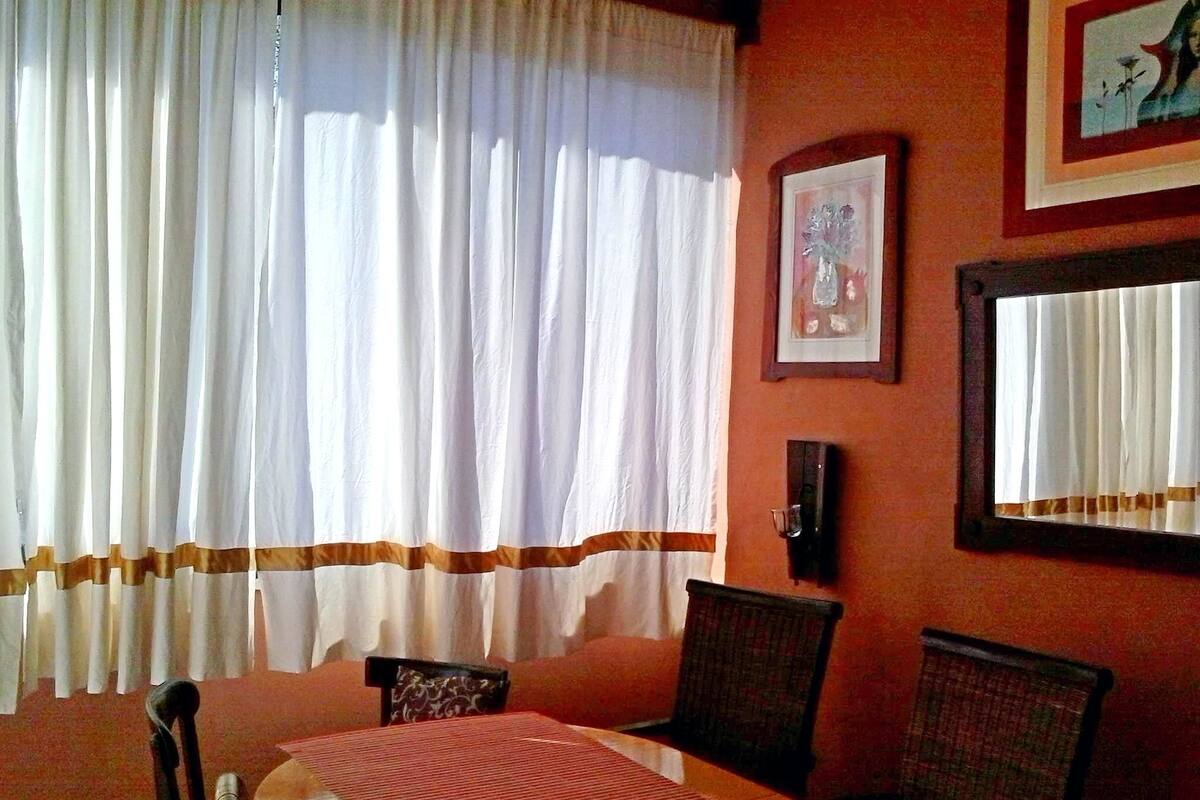 A dining area with a table set for four, chairs, a window with white curtains, and a framed picture on the wall.