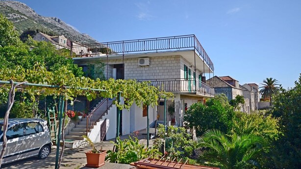 Een twee verdiepingen tellend huis met een balkon, omgeven door groen en een berg op de achtergrond.