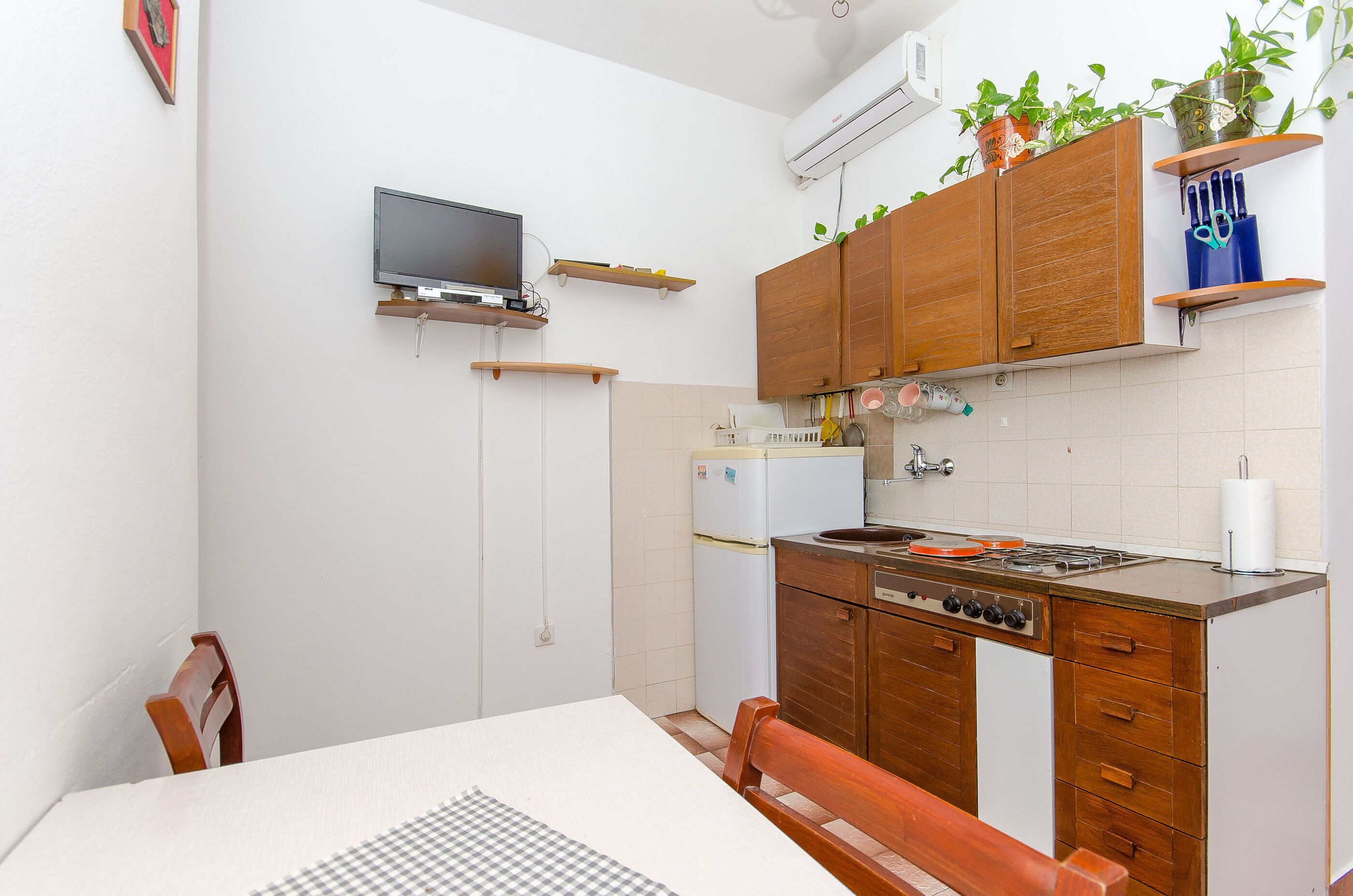 Una cocina compacta con armarios de madera, una estufa integrada, un refrigerador y un televisor montado en la pared.