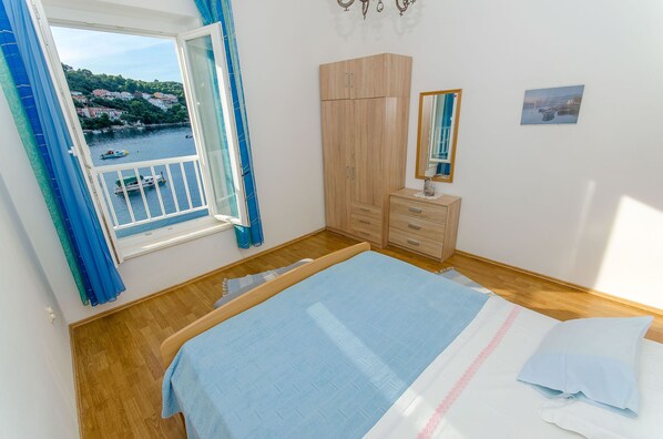 A bedroom with a bed, a wardrobe, a dresser, a window with a view of a body of water and buildings, and a mirror on the wall. - Apartment Jasenka (Brna)