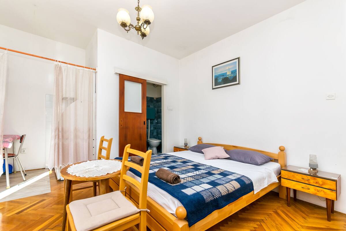 Een slaapkamer met een houten bed, een nachtkastje, een stoel, een klein tafeltje met een stoel, een kroonluchter en een schilderij aan de muur.