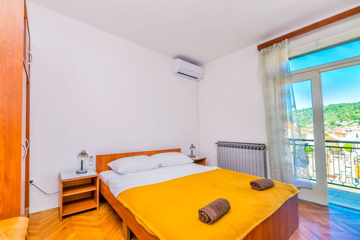 Een slaapkamer met een houten bed, witte kussens, een gele deken, een opgerolde handdoek, een nachtkastje met een lamp, en uitzicht op een balkon met een leuning.