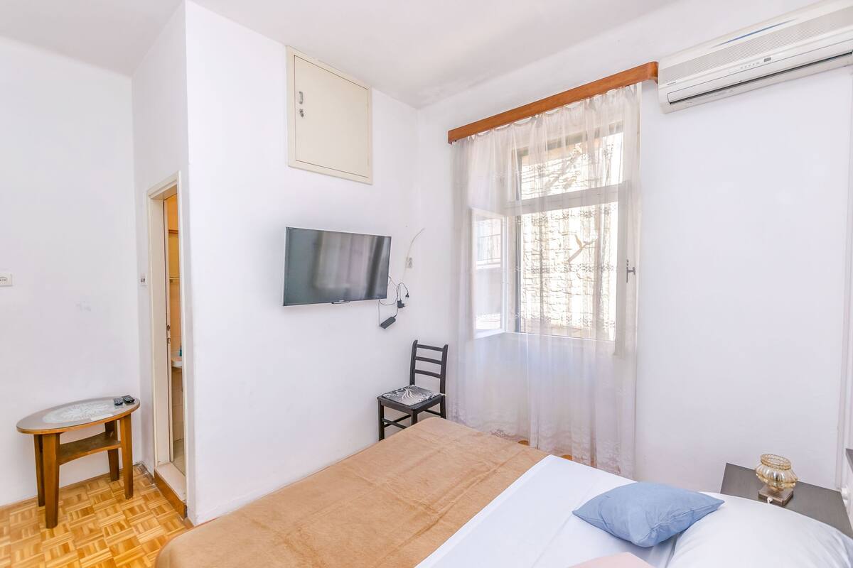 Een slaapkamer met een bed, een stoel, een aan de muur bevestigde televisie en een raam met gordijnen.